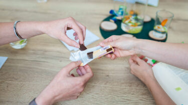 Eine Person bricht ein Stück Schokolade aus einer Verpackung, die ihr von einer anderen Person am Holztisch gereicht wird.