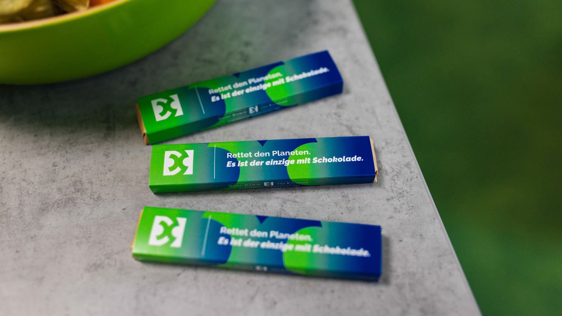 Drei bedruckte Schokoladentafeln von Dices-Domin & Kollegen mit dem Aufdruck „Rettet den Planeten. Es ist der einzige mit Schokolade.“ auf einem grauen Tisch.