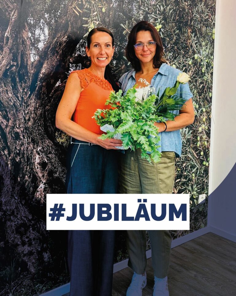 Zwei lächelnde Frauen stehen nebeneinander mit einem Blumentopf und einer Rose vor einer Olivenbaum-Fotowand mit dem Schriftzug #JUBILÄUM.