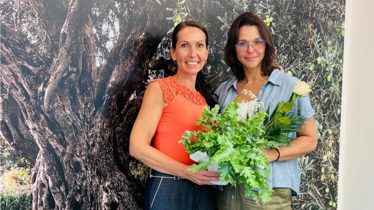 Zwei lächelnde Frauen stehen nebeneinander mit einem Blumentopf und einer Rose vor einer Olivenbaum-Fotowand mit dem Schriftzug #JUBILÄUM.
