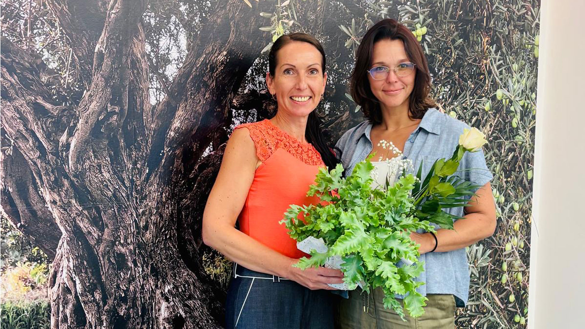 Zwei lächelnde Frauen stehen nebeneinander mit einem Blumentopf und einer Rose vor einer Olivenbaum-Fotowand mit dem Schriftzug #JUBILÄUM.